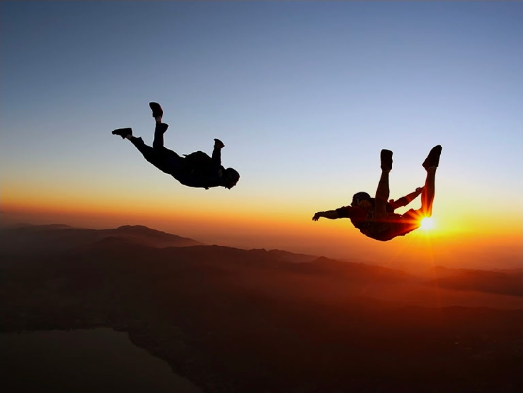 Skydivers silhouetted against a golden sunset above mountain ridges